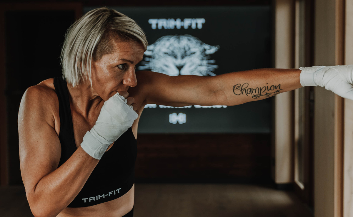 Frau mit kurzen blonden Haaren beim Boxen, zeigt ein Champion-Tattoo auf ihrem Arm und trägt Trainingskleidung.