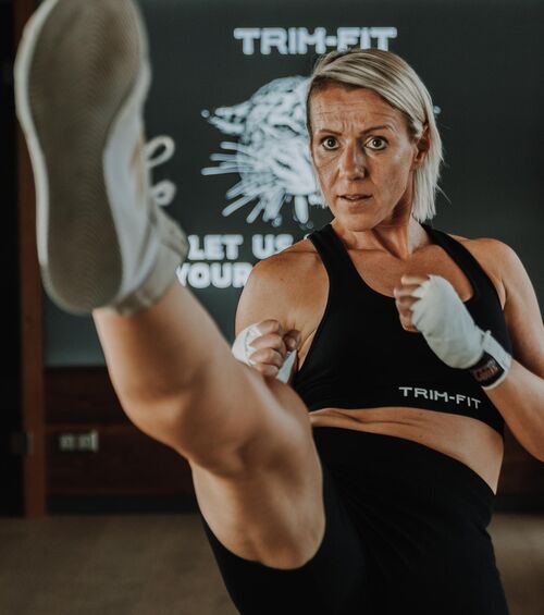 Eine Frau in Sportkleidung führt in einer Turnhalle einen hohen Tritt aus, ihr Gesichtsausdruck ist konzentriert.