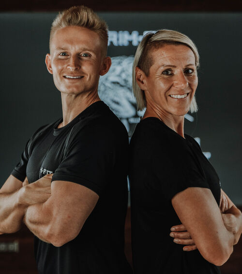 Zwei Personen stehen Rücken an Rücken, lächelnd, mit verschränkten Armen, beide tragen schwarze T-Shirts.