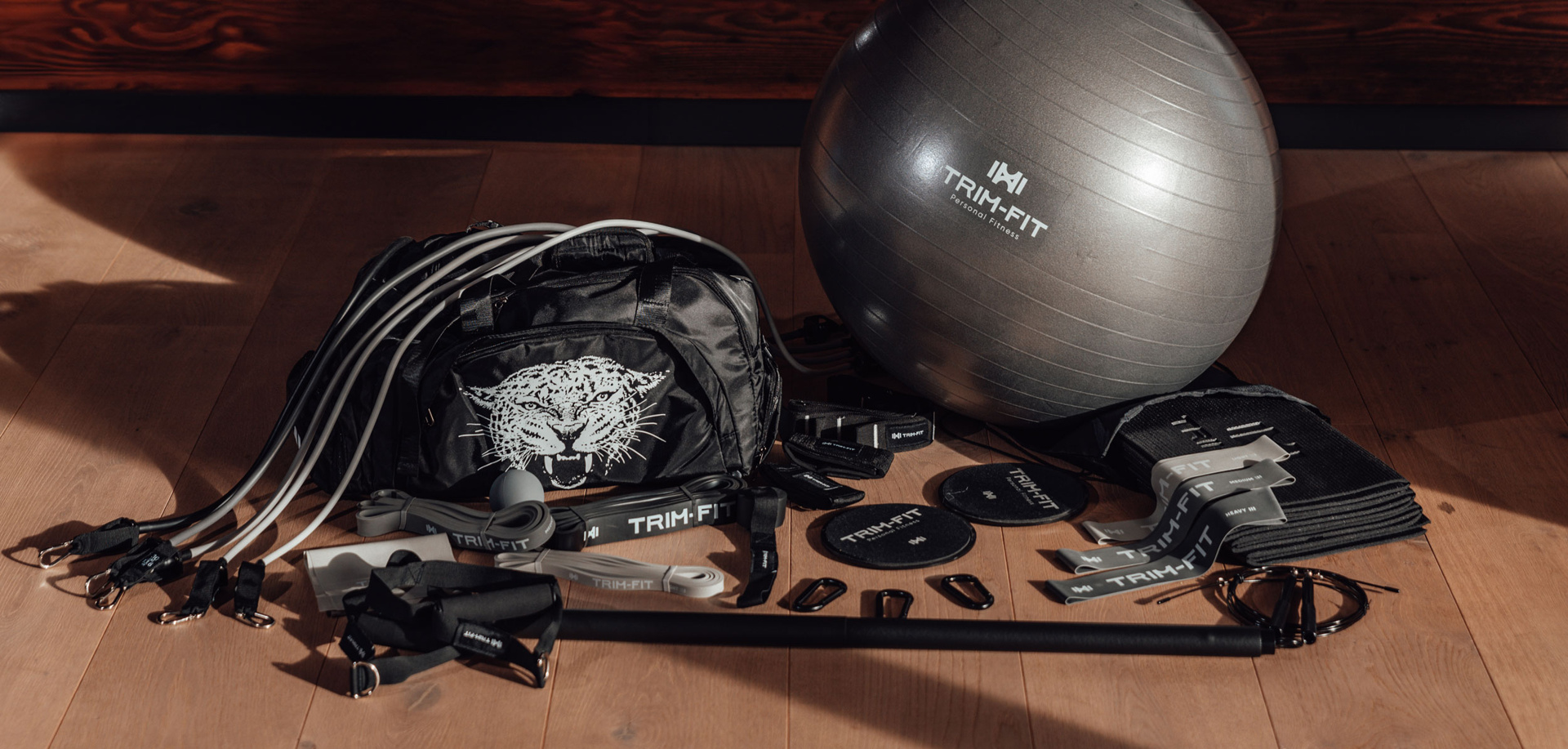 Verschiedene TRIBEFIT-Übungsgeräte, darunter Widerstandsbänder, ein Stabilitätsball und ein schwarzer Turnbeutel auf Holzboden.